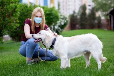 Koronavirüs salgını. Koruyucu maskeli genç bir kız yaz parkında köpeği gezdirir. Karantinada köpeği olan bir kadın.