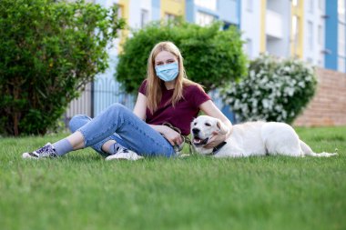 Koronavirüs salgını. Koruyucu maskeli genç bir kız yaz parkında köpeği gezdirir. Karantinada köpeği olan bir kadın.
