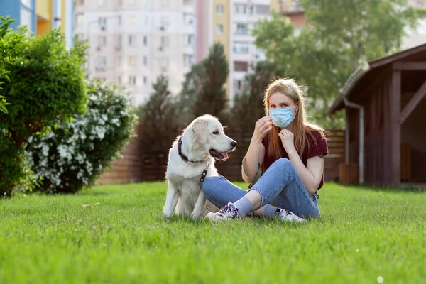Koronavirüs salgını. Koruyucu maskeli genç bir kız yaz parkında köpeği gezdirir. Karantinada köpeği olan bir kadın.