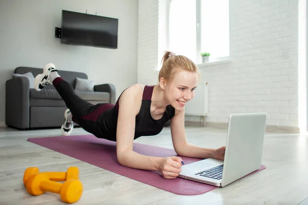 young sports girl go in for sports at home and watches video training on a laptop, athlete does exercise in the room in the morning and watches online tutorial, self-isolation concept
