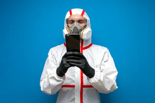 Coronavirus pandemic. disinfection worker in a protective suit and a respirator holds a phone and reads, a virologist on a blue isolated background