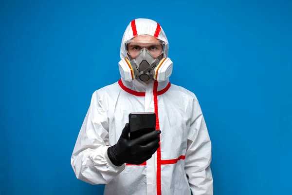 disinfection worker in a protective suit and respirator holds a phone and reads news about coronavirus, doctor virologist on a blue background