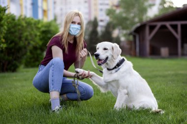 Coronavirüs salgınında köpeği gezdiren genç bir kız. Parkta köpek yavrusuyla maske takan bir kadın.