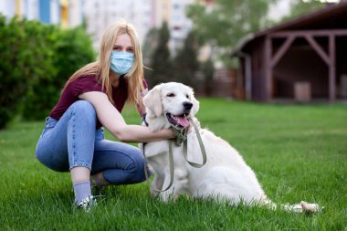 Koronavirüs salgını. Koruyucu maskeli genç bir kız yaz parkında köpeği gezdirir. Karantinada köpeği olan bir kadın.