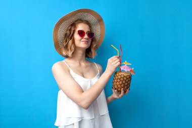 Yaz kıyafetleri içinde genç bir kız tropik bir ananas kokteyli içiyor ve mavi izole bir arka planda gülümsüyor. Egzotik bir içkiye sahip bir kadın bir otelde dinleniyor, deniz kenarında tatil kavramı.