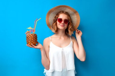 Tropik ananas kokteyli elbiseli genç bir kız mavi izole bir arka planda gülümsüyor, egzotik bir içkiye sahip bir kadın tatil köyünde dinleniyor, yaz tatili konsepti.