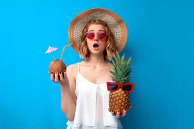 young shocked girl in summer clothes holds tropical cocktails made of pineapple and coconut on a blue isolated background, surprised woman at the resort with exotic drinks clipart