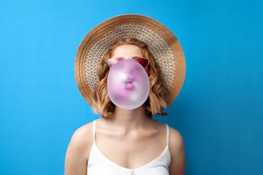 Güneş şapkalı ve gözlüklü genç bir kız mavi izole bir arka planda sakız şişiriyor. Bir kadın yaz kıyafetleri içinde pembe bir sakız çiğniyor.