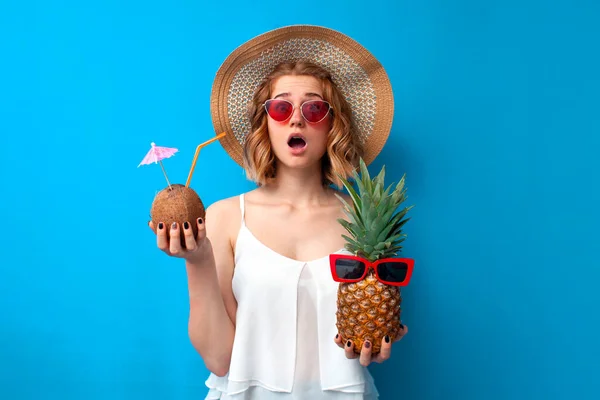 Yazlık elbiseli genç bir kız ananas ve hindistancevizinden yapılmış tropikal kokteylleri mavi izole bir arka planda tutuyor. Otelde egzotik içkilerle şaşırmış bir kadın.
