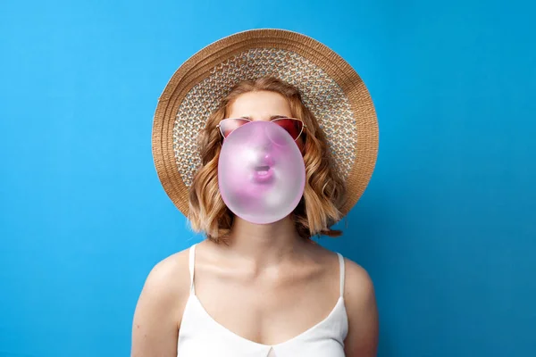 young girl in a sun hat and glasses inflates a bubble gum on a blue isolated background, a woman chews a pink chewing gum in summer clothes, close-up