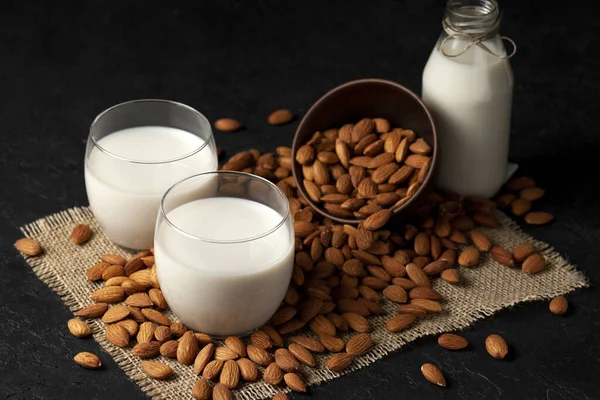 sugar-free and lactose-free almond milk in glasses and bottles with powdered nuts, vegetarian milk drink on a dark black table