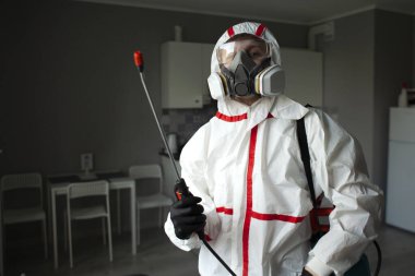 Haşere kontrolü. Koruyucu giysili bir işçi, arka planda spreyli bir apartman dairesinde çalışıyor. Sağlık hizmetleri odayı kimyasal bir maddeyle dezenfekte ediyor.