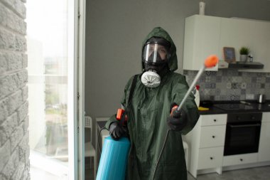 Haşere kontrolü. Koruyucu kıyafetli bir işçi mutfaktaki rafları hamamböcekleri ve karıncalardan spreyle temizler. Sağlık servisi odayı kimyasal bir maddeyle dezenfekte eder.
