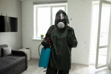 Haşere kontrolü. Koruyucu giysili bir işçi, arka planda spreyli bir apartman dairesinde çalışıyor. Sağlık hizmetleri odayı kimyasal bir maddeyle dezenfekte ediyor.