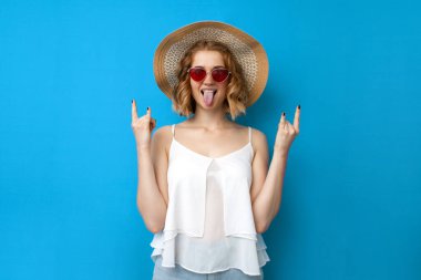 Yaz konsepti. Güneş şapkalı ve gözlüklü genç çılgın kız mavi izole bir arka planda rocknrol işareti, yaz kıyafetleri içinde tatil yapan bir kadın gösteriyor.