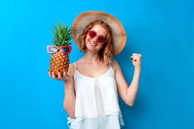 Genç çılgın kız, güneş şapkası ve gözlükleriyle mavi izole bir arka planda ananas, tatil köyünde kadın turist ve yaz tatili konseptiyle zafere seviniyor.