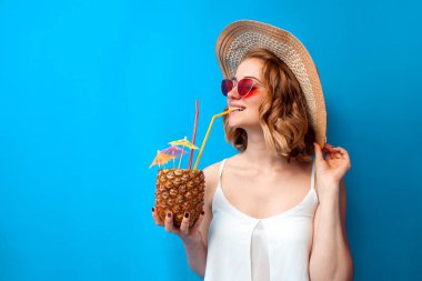 Yaz kıyafetleri içinde genç bir kız tropik bir ananas kokteyli içiyor ve mavi izole bir arka planda gülümsüyor. Egzotik bir içkiye sahip bir kadın bir otelde dinleniyor, deniz kenarında tatil kavramı.