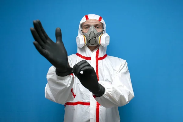 worker in a chemical protective suit puts on gloves on a blue isolated background, health worker