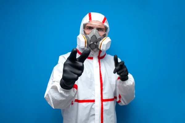 man in a chemical protective suit shows his fingers at you on a blue isolated background, an epidemiologist, biologist, virologist, scientist