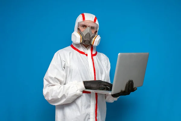 man in protective chemical suit holds a laptop on a blue isolated background, doctor virologist use computer