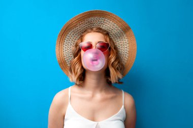 Güneş şapkalı ve gözlüklü genç bir kız mavi izole bir arka planda sakız şişiriyor. Bir kadın yaz kıyafetleri içinde pembe bir sakız çiğniyor.