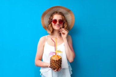 Güneş şapkalı ve gözlüklü genç kız tropikal bir ananas kokteyli tutuyor ve mavi izole bir arka planda gülümsüyor. Egzotik içkisi olan bir kadın tatil köyünde dinleniyor, yaz konseptinde.