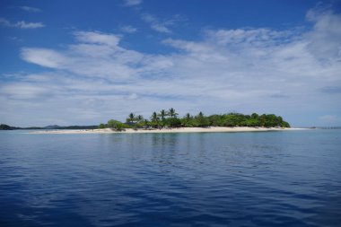Güneşli bir günde çok sakin bir denizin görüntüsü. Gökyüzünde birkaç dağınık bulut var. Ufukta küçük bir ada görüyoruz. Palmiye ağaçları ve davetkar, kumlu bir plaj.. 