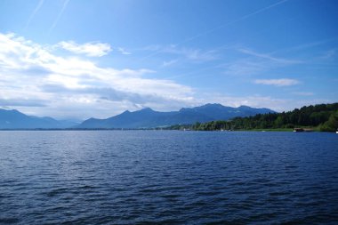 Chiemsee Gölü 'nün (Bavyera, Almanya) manzarası. Uzakta dağların yanı sıra orman adaları görülebilir. Gökyüzü çok mavi, dağınık bulutlar var. Hafif dalgalı deniz tarağı suları.