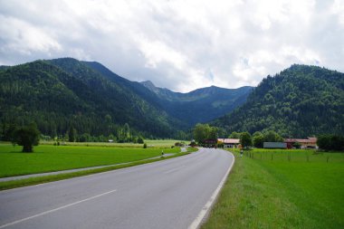 Dağ köyüne doğru giden yeşil tarlalardan geçen boş bir asfalt yol. Uzakta, yoğun ormanlık yeşil dağlar ve birkaç bina var. Gökyüzü çok bulutlu..