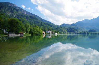 Orman yeşili dağlarla çevrili güzel mavi gölü olan bir manzara. Uzakta ağaçların arasına gizlenmiş birkaç bina var. Gökyüzünde bulutlar olan aydınlık bir yaz günü..