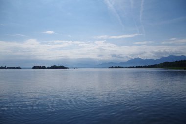 Chiemsee Gölü 'nün (Bavyera, Almanya) manzarası. Uzakta dağların yanı sıra orman adaları görülebilir. Gökyüzü çok mavi, dağınık bulutlar var. Hafif dalgalı deniz tarağı suları.