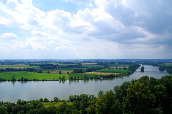 A landscape with the Danube river, viewed from Walhalla (Germany). Green and yellow fields dominate. Thick grove of trees in the foreground. Very distant towns can be seen. Dark clouds in the sky.