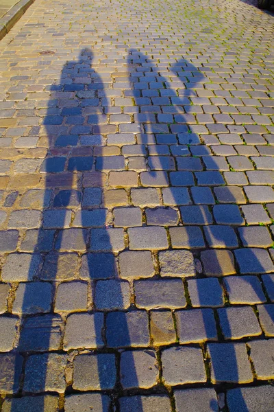 Dark, elongated shadows of man and woman on an old cobblestone pavement on a sunny day. Stones are colorful and reflect the sun rays.