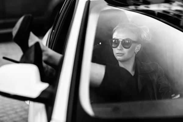 Photo de mode en plein air de belle femme sexy aux cheveux foncés en veste en cuir noir et lunettes de soleil posant en voiture de luxe — Photo