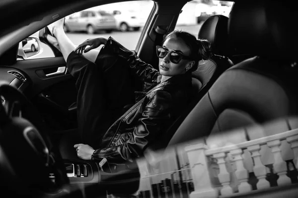 Photo de mode en plein air de belle femme sexy aux cheveux foncés en veste en cuir noir et lunettes de soleil posant en voiture de luxe — Photo