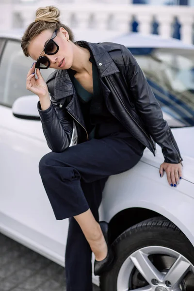 Photo de mode en plein air de belle femme sexy aux cheveux foncés en veste en cuir noir et lunettes de soleil posant en voiture de luxe — Photo