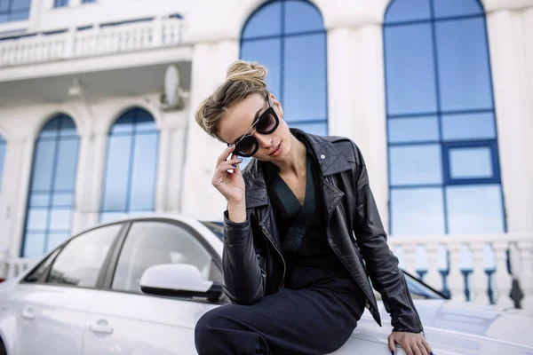 Foto al aire libre de moda de mujer hermosa sexy con pelo oscuro en chaqueta de cuero negro y gafas de sol posando en auto de lujo — Foto de Stock