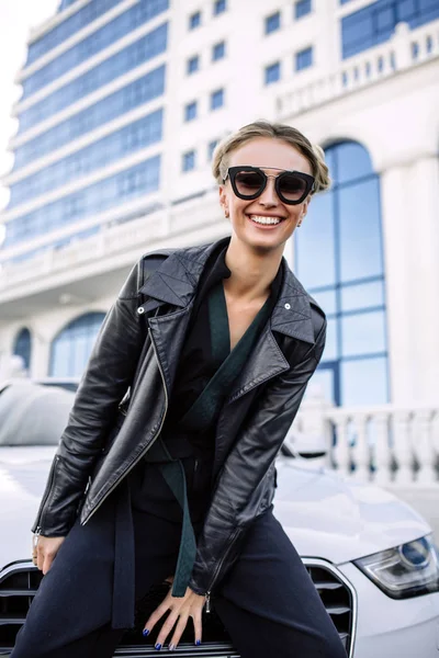 Photo de mode en plein air de belle femme sexy aux cheveux foncés en veste en cuir noir et lunettes de soleil posant en voiture de luxe — Photo