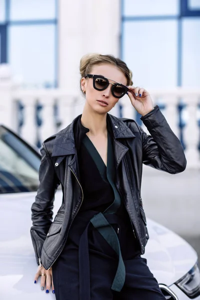 Foto al aire libre de moda de mujer hermosa sexy con pelo oscuro en chaqueta de cuero negro y gafas de sol posando en auto de lujo — Foto de Stock
