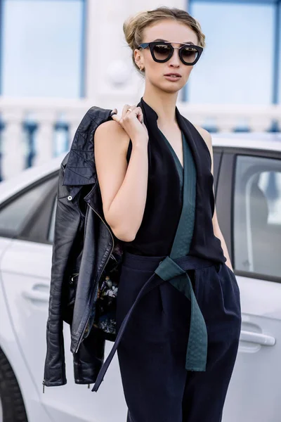 Foto al aire libre de moda de mujer hermosa sexy con pelo oscuro en chaqueta de cuero negro y gafas de sol posando en auto de lujo — Foto de Stock