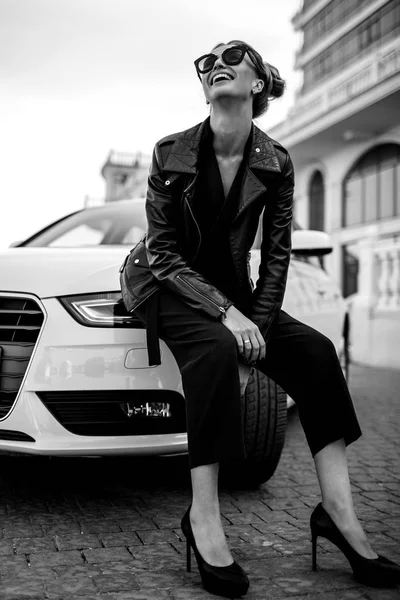 Photo de mode en plein air de belle femme sexy aux cheveux foncés en veste en cuir noir et lunettes de soleil posant en voiture de luxe — Photo