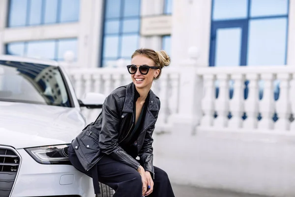 Photo de mode en plein air de belle femme sexy aux cheveux foncés en veste en cuir noir et lunettes de soleil posant en voiture de luxe — Photo