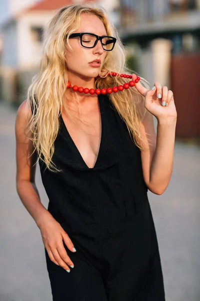 Porträtt av en flicka blondin i en svart klänning ögon glasögon på halsen av röda pärlor. Håller sin högra hand finger pärlor. — Stockfoto