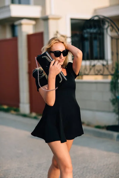 Porträtt av vacker blond flicka i svarta glasögon och svart klänning medföljer handväska. — Stockfoto