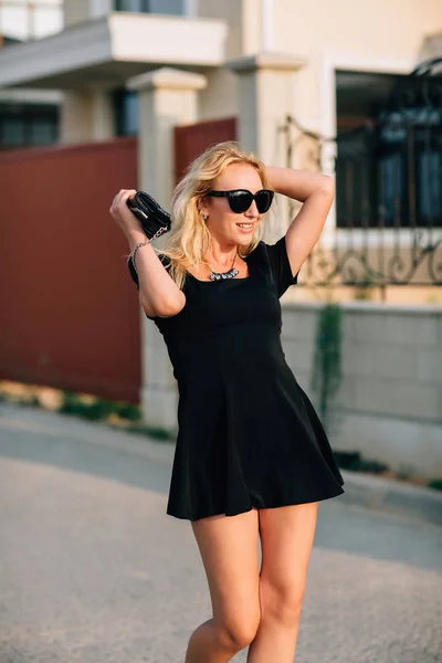 Портрет красивой блондинки в черных очках и черном платье поставляется с сумочкой . — стоковое фото
