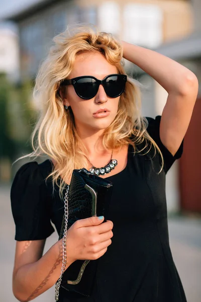 Porträtt av vacker blond flicka i svarta glasögon och svart klänning medföljer handväska. — Stockfoto