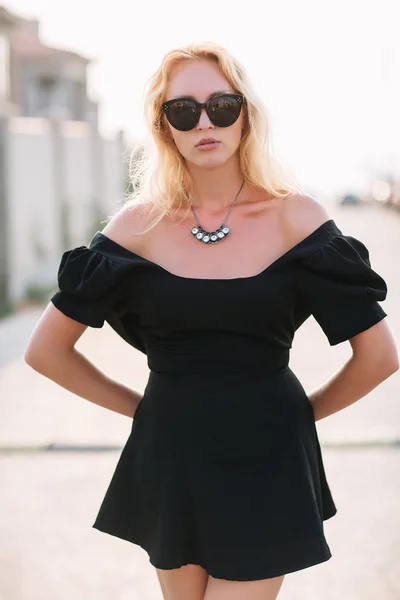 Portrét krásné blond dívka v černé brýle a černé šaty přichází s kabelka. — Stock fotografie