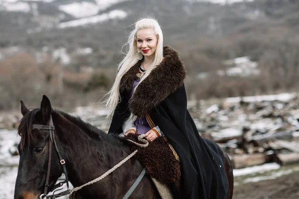 Hermosa rubia vikinga en una capa negra a caballo — Foto de Stock