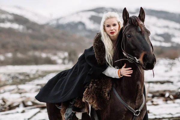 Beautiful blonde Viking in a black cape on horseback