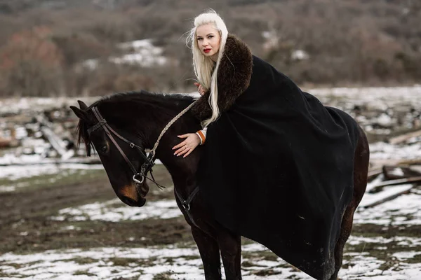 Vacker blondin Viking i en svart kappa på hästryggen — Stockfoto
